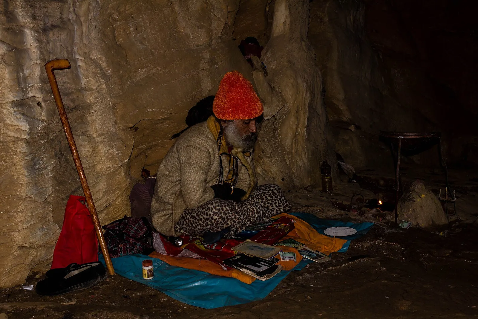 A sadhu in an orange woollen hat seated on a mat inside the rock entrance of Siddha Gufa