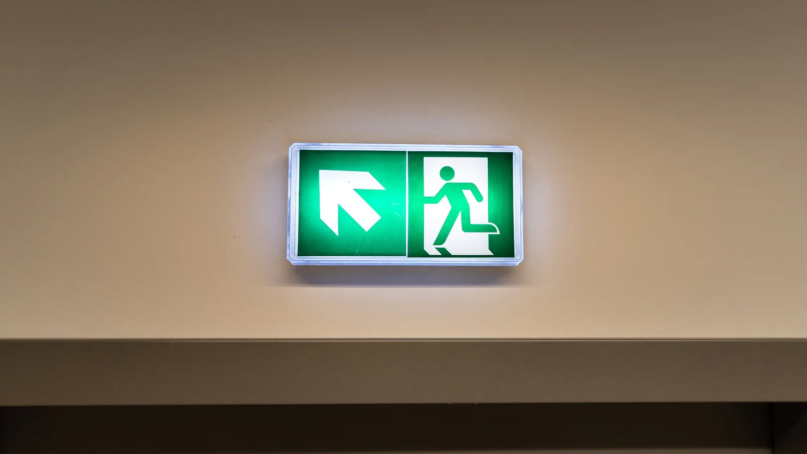 A green emergency exit sign with an arrow on a pale wall