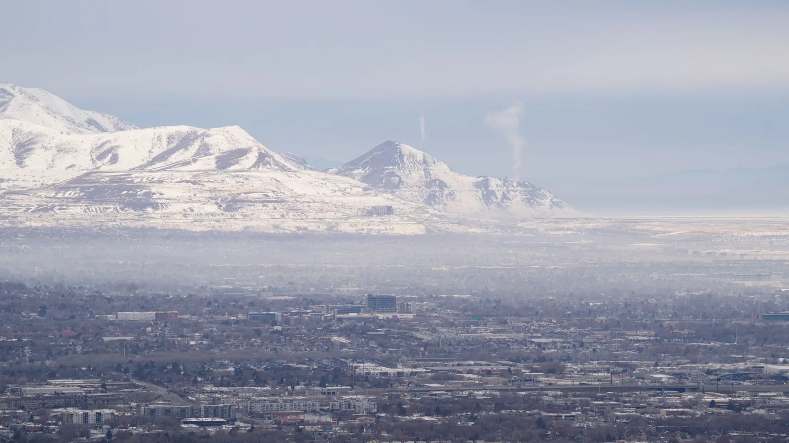 Winter air pollution and haze fill a valley beneath snow-capped mountains, illustrating the soot-laden smog that settles onto high-altitude snow and darkens it