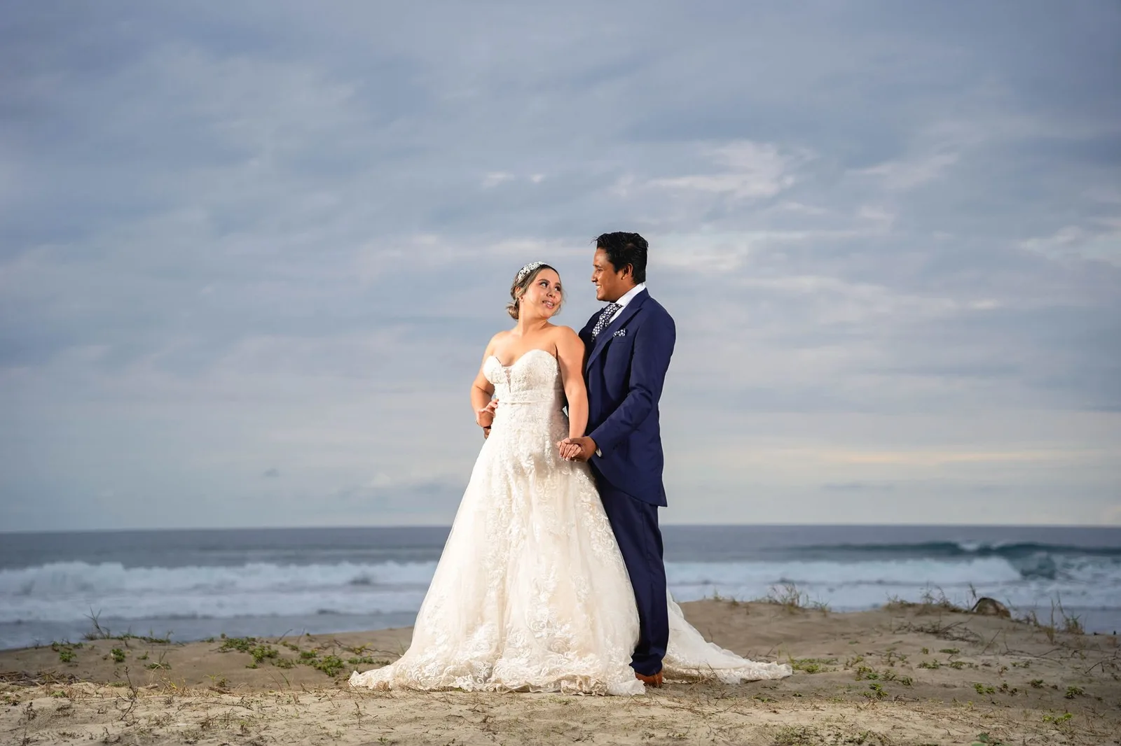 A newlywed couple in wedding dress and suit walking hand in hand on a beach at a destination-wedding honeymoon