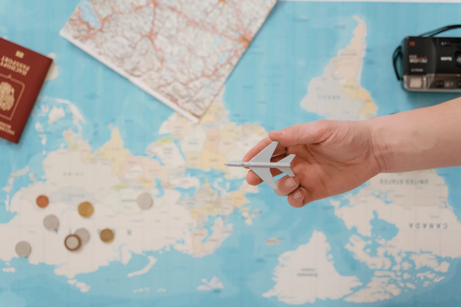 A hand holds a small toy aeroplane over a world map surrounded by coins, a camera and a passport — planning the budget for a year of travel.