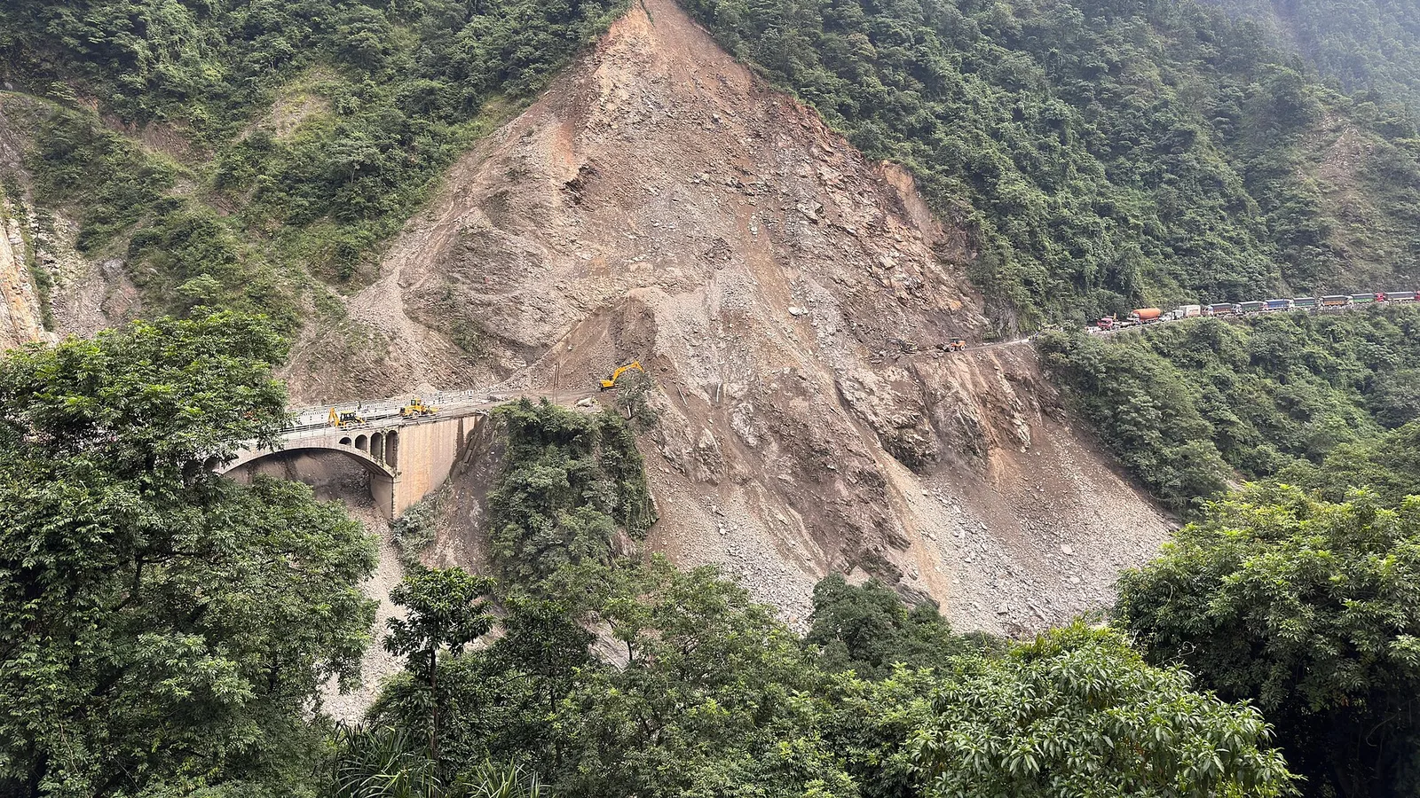 A large landslide scar tears down a steep, forested hillside onto a mountain highway in Nepal, where excavators clear the debris and a line of trucks waits beyond the blocked road