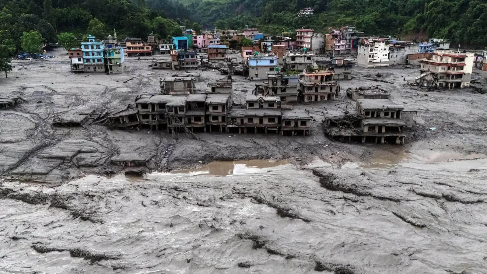 The Trishuli riverbank at Nuwakot after the 26 August 2026 flood, a row of gutted buildings half-buried in grey silt with an intact hillside settlement above them. Photograph shared on Instagram.