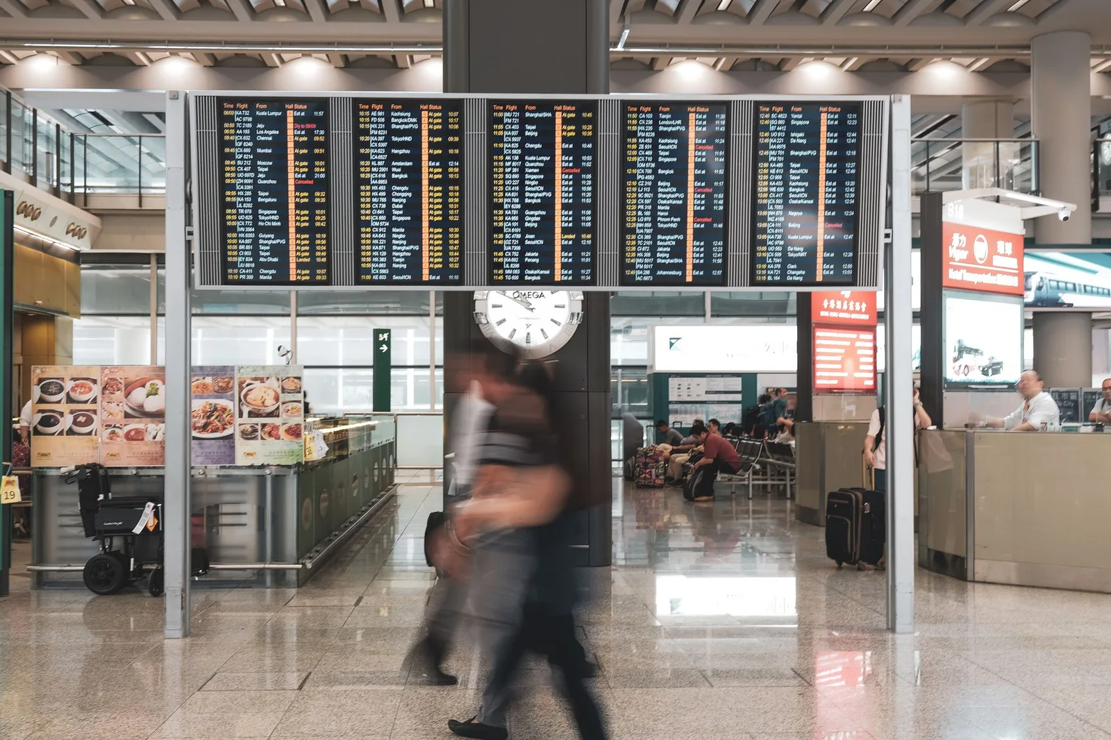 A departure board showing delayed flights while travellers wait in an airport terminal