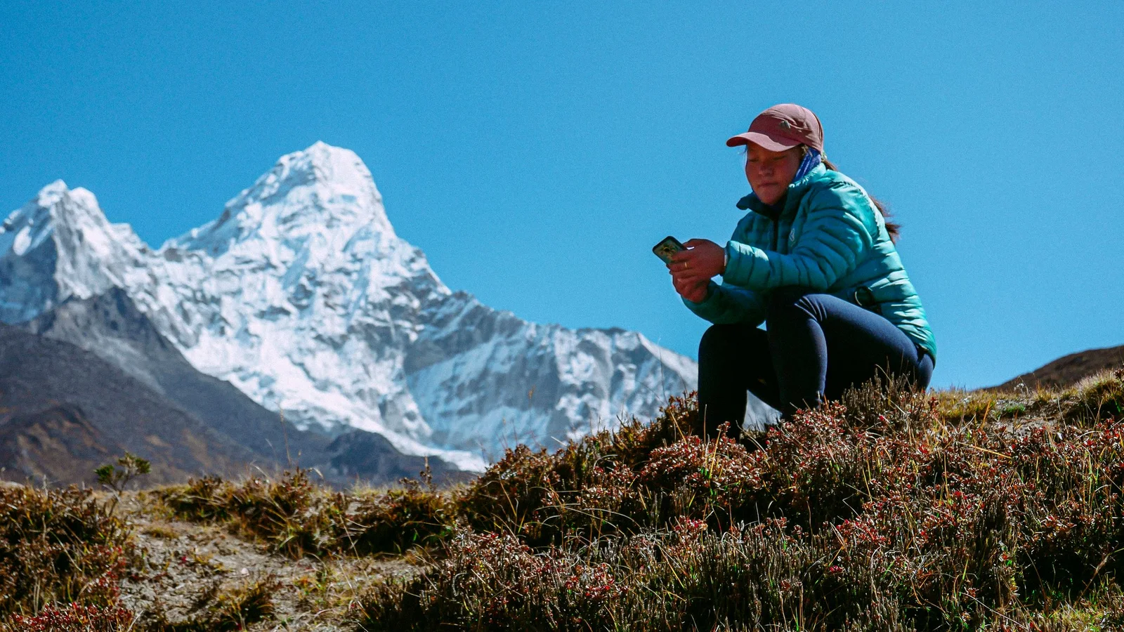 Woman sits on a hilltop using phone with Ama Dablam mountain backdrop in Nepal.