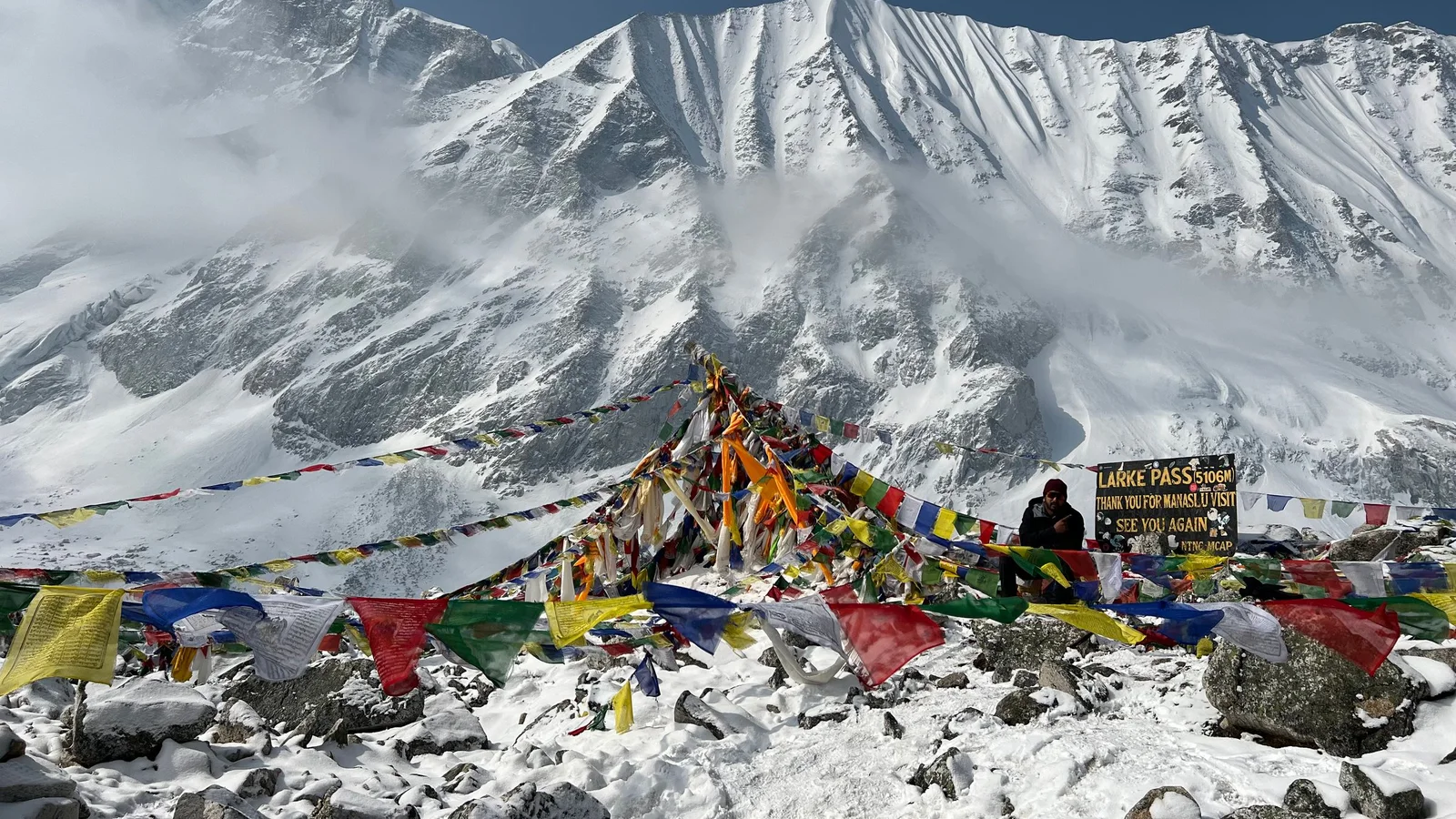 Colorful prayer flags flutter at snow-covered Larke Pass under majestic mountains.