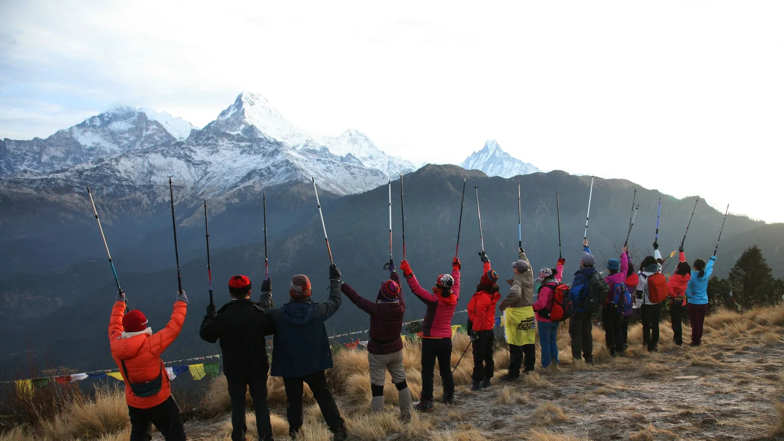 Hikers raise trekking poles in celebration with the Himalayan mountains in the background, a symbol of adventure and achievement.