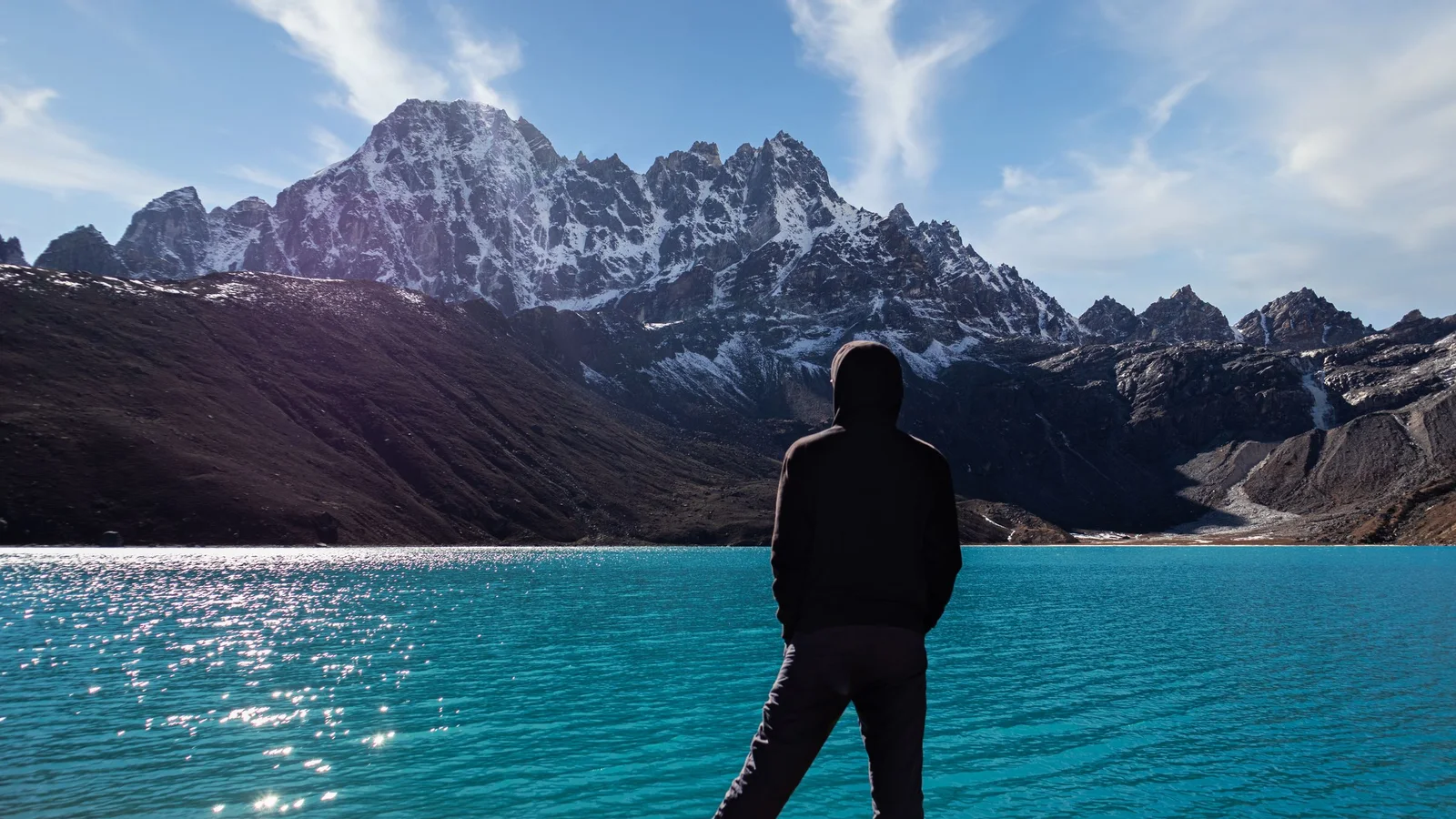 A lone figure in a hoodie gazes at a turquoise lake with majestic snow-capped mountains.