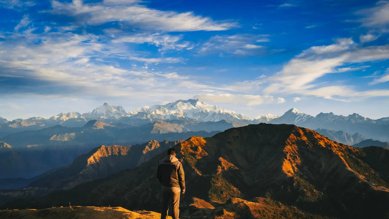 A person stands on a mountain peak, overlooking a stunning sunrise over the Himalayas.